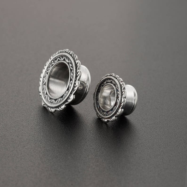 May include: Two silver ear gauges with an ornate design. The larger gauge has a diameter of approximately 1 inch, and the smaller gauge has a diameter of approximately 1/2 inch.