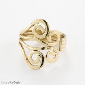 Pode incluir: Um anel dourado com um design único com padrões redemoinhantes e vazados. O anel tem uma superfície texturizada e parece ser ajustável. O anel está sobre um fundo branco.