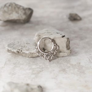 K&ouml;nnte beinhalten: Ein silberner Septumring mit einem dekorativen, verzierten Design. Der Ring ist aus Metall und hat eine kreisf&ouml;rmige Form mit einem baumelnden, komplizierten Muster.