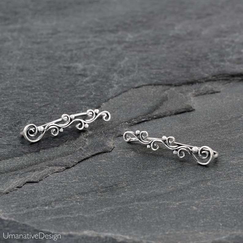 May include: A pair of silver ear cuffs with a delicate, floral design. The cuffs are made of a thin, twisted wire and feature small, round beads.
