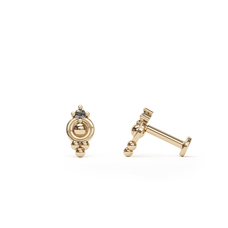 May include: A gold body jewelry piece with a small black gemstone and a cluster of gold beads. The jewelry is designed for a cartilage piercing.