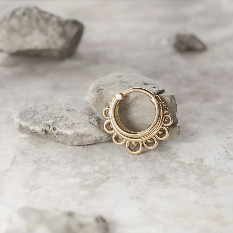 May include: Gold septum ring with a decorative design of multiple circles. The ring is on a gray rock.