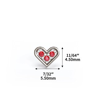 Puede incluir: Un amuleto de plata en forma de coraz&oacute;n con piedras preciosas rojas y blancas. El amuleto mide 5,50 mm de ancho y 4,50 mm de alto.