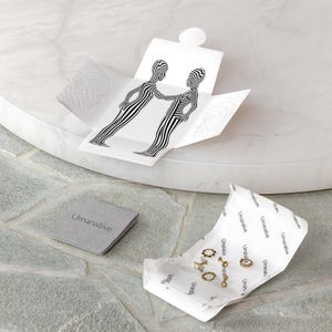 May include: A set of gold hoop earrings and a gold chain necklace with a small pendant, packaged in a white box with a black and white graphic design. The box is open and the jewelry is visible. The jewelry is on a white paper with the word "Umanative" printed on it.