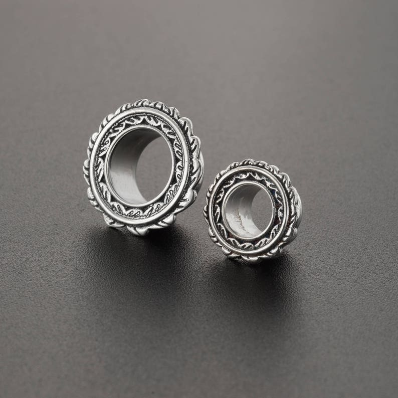 May include: Two silver ear gauges with intricate, ornate designs. The larger gauge has a diameter of approximately 1 inch, while the smaller gauge is about 0.75 inches in diameter.