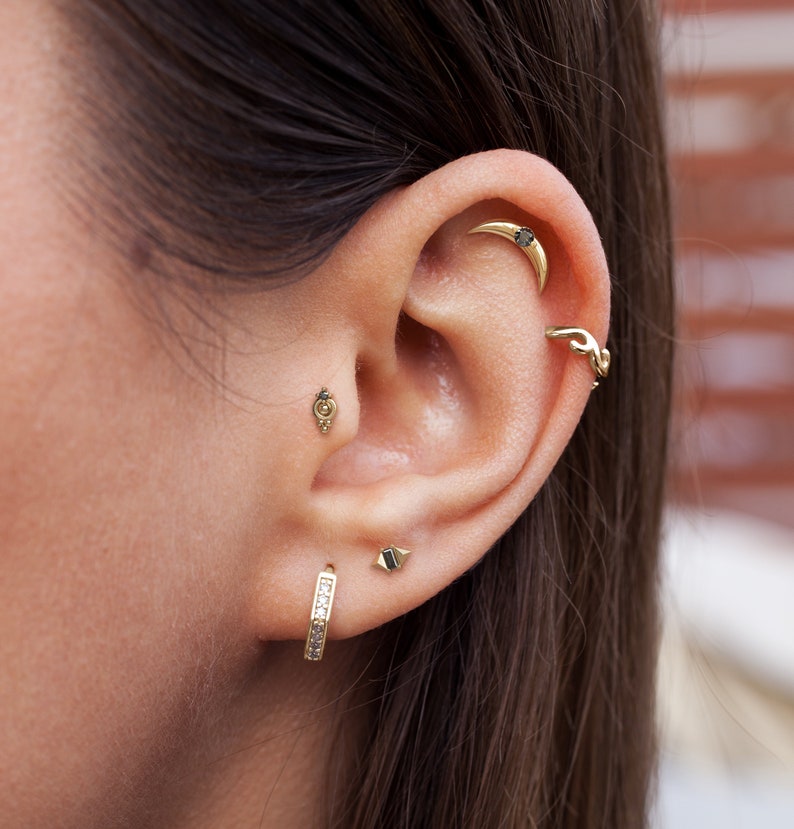 May include: A close-up of a person's ear with multiple piercings. The ear is pierced with a hoop earring, a small stud, a curved bar, and a small hoop. The piercings are all adorned with gold jewelry.
