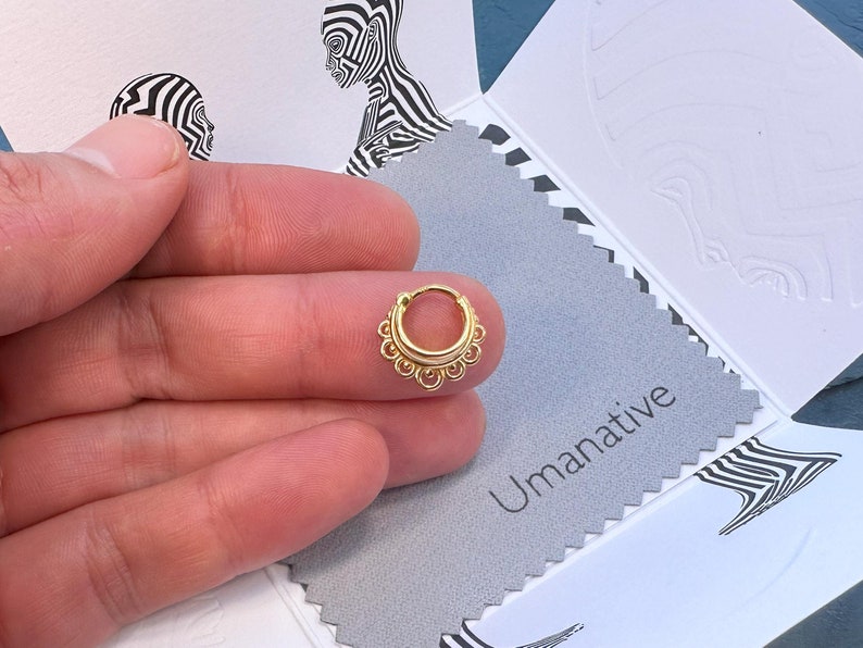May include: Gold hoop nose ring with a decorative design around the hoop. The ring is on a finger.