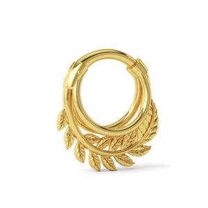 Puede incluir: Un anillo de tabique dorado con un diseño de hojas. El anillo circular tiene un cierre de bisagra y un elemento decorativo de hojas estilizadas. La joya está hecha de un metal brillante y reflectante.