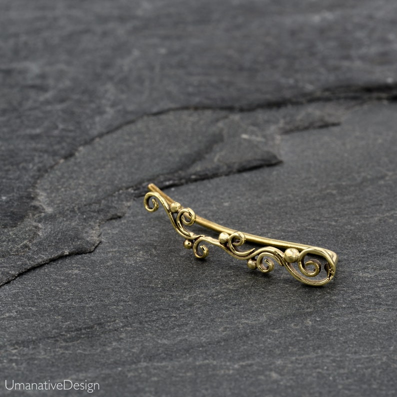 May include: A gold-toned ear cuff with a delicate, ornate design featuring swirling lines and small beads. The cuff is curved and has a slightly asymmetrical shape.
