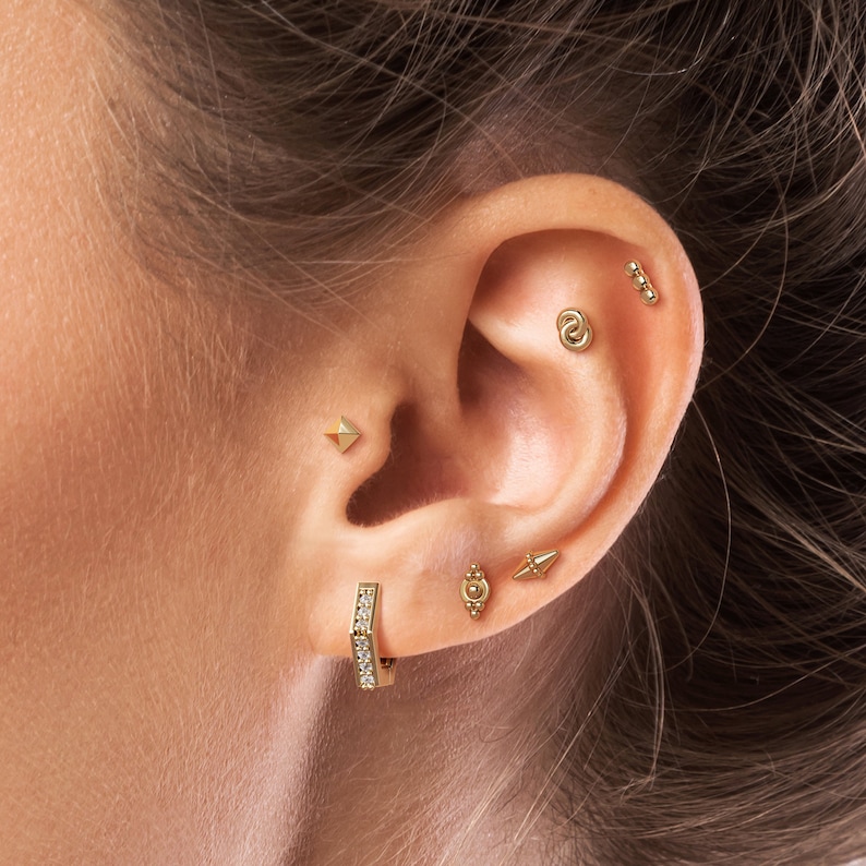 May include: A close-up of a person's ear with multiple gold earrings. The earrings include a small diamond hoop, a small triangle stud, a small circle stud, a small bar stud with three beads, and a small stud with a circle and a small triangle.