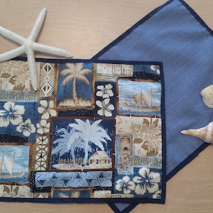 May include: A blue and brown placemat with a tropical design featuring palm trees, flowers, and sailboats. The placemat has a dark blue border and is made of fabric.