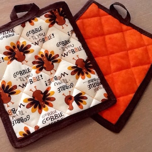 May include: Two quilted pot holders. One is orange with a brown border. The other is white with a brown border and a turkey pattern. The pattern includes the text "Gobble Til You Wobble!"