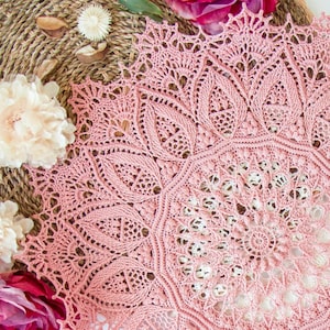 May include: A delicate pink crochet doily with a lacy design. The doily is circular and has a centre that is slightly lighter in colour than the outer edges.