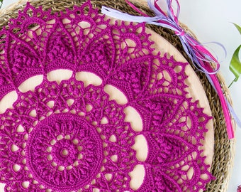 Può includere: Un grande tovagliolo all'uncinetto magenta con un design dettagliato e intricato. Il tovagliolo è appoggiato su una base circolare intrecciata.