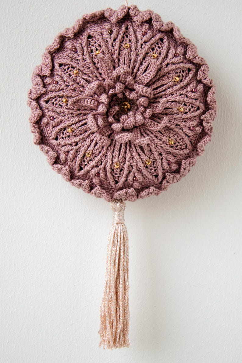 Puede incluir: Un adorno de pared redondo, hecho a mano, en color rosa empolvado. La pieza presenta un intrincado trabajo de ganchillo con un dise&ntilde;o floral, adornado con peque&ntilde;as cuentas doradas. Una borla a juego cuelga de la parte inferior, a&ntilde;adiendo un elemento decorativo.