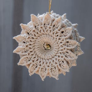 Puede incluir: Adorno beige tejido a ganchillo con dise&ntilde;o floral. El adorno redondo tiene una cuenta dorada central y un hilo dorado para colgar. El tejido a ganchillo presenta intrincados patrones calados.