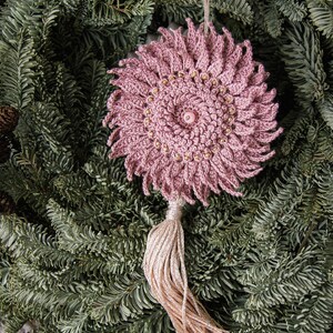 Puede incluir: Adorno de ganchillo rosa polvoriento hecho a mano con un dise&ntilde;o floral. El adorno presenta un bot&oacute;n central y est&aacute; adornado con peque&ntilde;as cuentas doradas. Una borla a juego cuelga debajo. El adorno se exhibe sobre agujas de pino verdes.