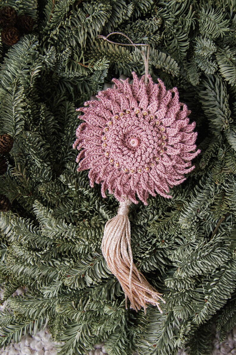 Puede incluir: Adorno de ganchillo rosa polvoriento hecho a mano con un dise&ntilde;o floral. El adorno presenta un bot&oacute;n central y est&aacute; adornado con peque&ntilde;as cuentas doradas. Una borla a juego cuelga debajo. El adorno se exhibe sobre agujas de pino verdes.