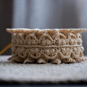 Puede incluir: Un recipiente beige tejido a ganchillo con un borde festoneado y una banda decorativa. La pieza hecha a mano es cil&iacute;ndrica y se asienta sobre una superficie texturizada. El hilo es de color beige claro.