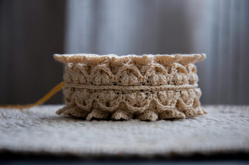 Puede incluir: Un recipiente beige tejido a ganchillo con un borde festoneado y una banda decorativa. La pieza hecha a mano es cil&iacute;ndrica y se asienta sobre una superficie texturizada. El hilo es de color beige claro.