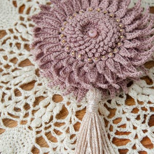 Puede incluir: Una flor de ganchillo hecha a mano en rosa polvoriento con cuentas doradas y una cuenta central rosa. La flor tiene una borla y est&aacute; colocada sobre un tapete de encaje color crema. Es un art&iacute;culo decorativo.
