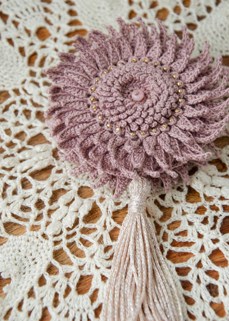 Puede incluir: Una flor de ganchillo hecha a mano en rosa polvoriento con cuentas doradas y una cuenta central rosa. La flor tiene una borla y est&aacute; colocada sobre un tapete de encaje color crema. Es un art&iacute;culo decorativo.