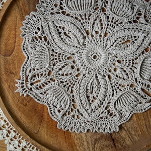Può includere: Un centrino all'uncinetto fatto a mano, bianco sporco, con un motivo floreale, appoggiato su un vassoio di legno. Il centrino presenta un'intricata lavorazione traforata e un bordo smerlato. Un piccolo bordo in pizzo bianco è visibile a sinistra.