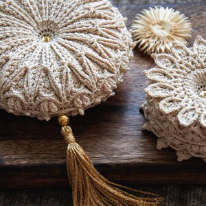 Puede incluir: Primer plano de art&iacute;culos de ganchillo beige, que incluyen un coj&iacute;n redondo con una borla dorada y una pila de posavasos. El coj&iacute;n tiene una cuenta dorada central y patrones intrincados. Los posavasos tienen forma de flor.