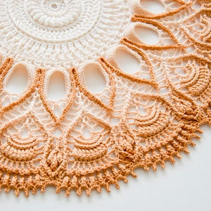 May include: Close-up of a handmade, circular crochet doily. The doily features a detailed pattern with a gradient of colors, transitioning from white to peach and then to a deeper orange-brown. The edges are scalloped, adding a decorative touch.