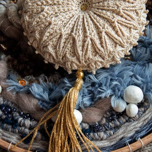 Puede incluir: Un coj&iacute;n redondo beige tejido a ganchillo con borla y bot&oacute;n dorados. El coj&iacute;n presenta un dise&ntilde;o texturizado y se asienta sobre un textil tejido con elementos azules, marrones y grises. Tambi&eacute;n hay cuentas decorativas y peque&ntilde;as esferas blancas.