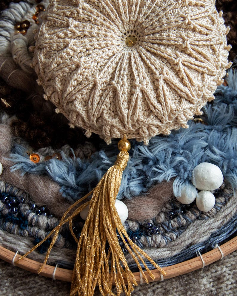 Puede incluir: Un coj&iacute;n redondo beige tejido a ganchillo con borla y bot&oacute;n dorados. El coj&iacute;n presenta un dise&ntilde;o texturizado y se asienta sobre un textil tejido con elementos azules, marrones y grises. Tambi&eacute;n hay cuentas decorativas y peque&ntilde;as esferas blancas.