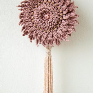 Puede incluir: Colgante de pared de ganchillo rosa empolvado hecho a mano, con dise&ntilde;o floral. El centro presenta una cuenta rosa, rodeada de cuentas doradas. Una larga borla beige cuelga debajo. Decoraci&oacute;n bohemia.