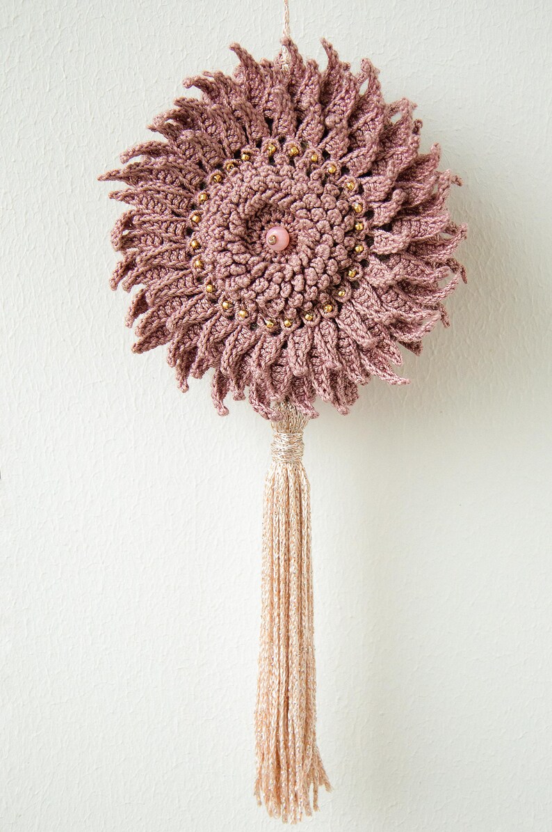 Puede incluir: Colgante de pared de ganchillo rosa empolvado hecho a mano, con dise&ntilde;o floral. El centro presenta una cuenta rosa, rodeada de cuentas doradas. Una larga borla beige cuelga debajo. Decoraci&oacute;n bohemia.