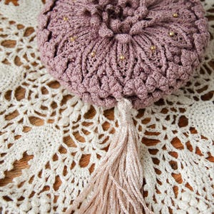 Puede incluir: Una bolsa de ganchillo rosa polvoriento con una borla, adornada con peque&ntilde;as cuentas doradas. La bolsa es redonda con un patr&oacute;n texturizado en forma de flor. La borla es de color beige claro. La bolsa descansa sobre un tapete de ganchillo blanco.