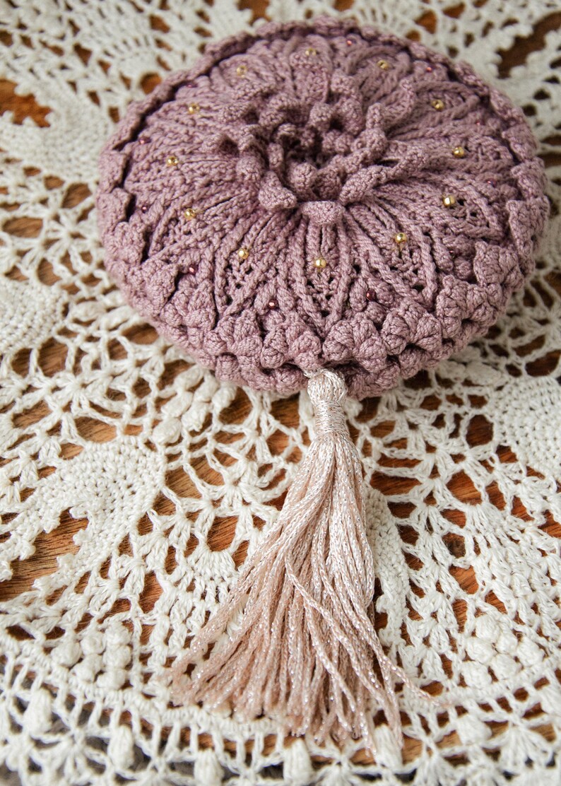 Puede incluir: Una bolsa de ganchillo rosa polvoriento con una borla, adornada con peque&ntilde;as cuentas doradas. La bolsa es redonda con un patr&oacute;n texturizado en forma de flor. La borla es de color beige claro. La bolsa descansa sobre un tapete de ganchillo blanco.