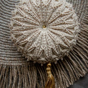 May include: A beige, round, crocheted ornament with a gold tassel. The ornament features a detailed, textured pattern and a small gold bead in the centre. The tassel is made of gold thread and hangs from the bottom.