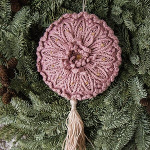 Puede incluir: Adorno de ganchillo rosa empolvado hecho a mano, con dise&ntilde;o floral y detalles de cuentas doradas. El adorno tiene una borla y se muestra sobre ramas de hoja perenne, ideal para la decoraci&oacute;n navide&ntilde;a.