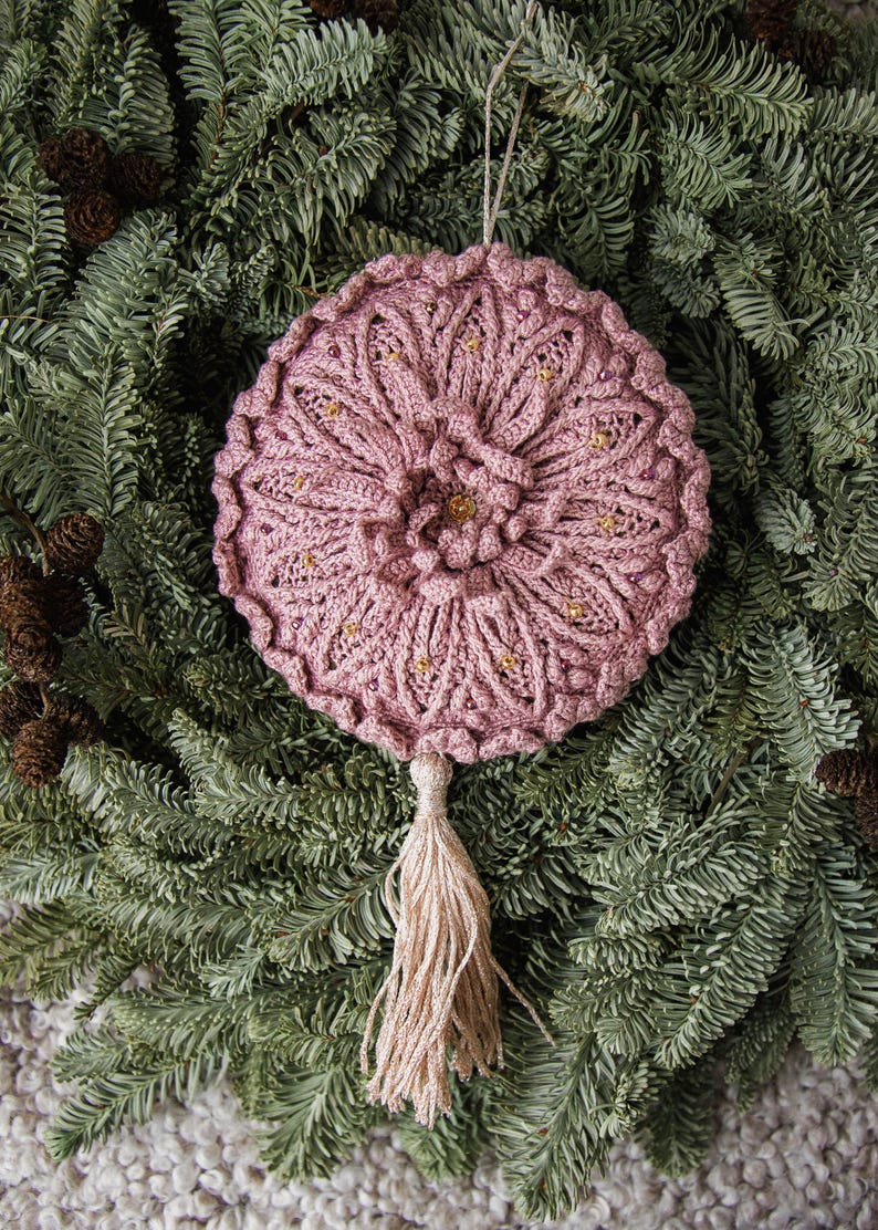 Puede incluir: Adorno de ganchillo rosa empolvado hecho a mano, con dise&ntilde;o floral y detalles de cuentas doradas. El adorno tiene una borla y se muestra sobre ramas de hoja perenne, ideal para la decoraci&oacute;n navide&ntilde;a.