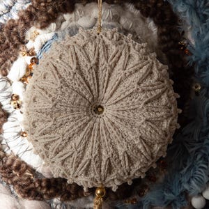 Puede incluir: Adorno beige tejido a ganchillo con dise&ntilde;o de estrella. El adorno redondo tiene una cuerda dorada para colgar y una borla. El fondo presenta varias texturas y colores, incluyendo azul, marr&oacute;n y blanco.