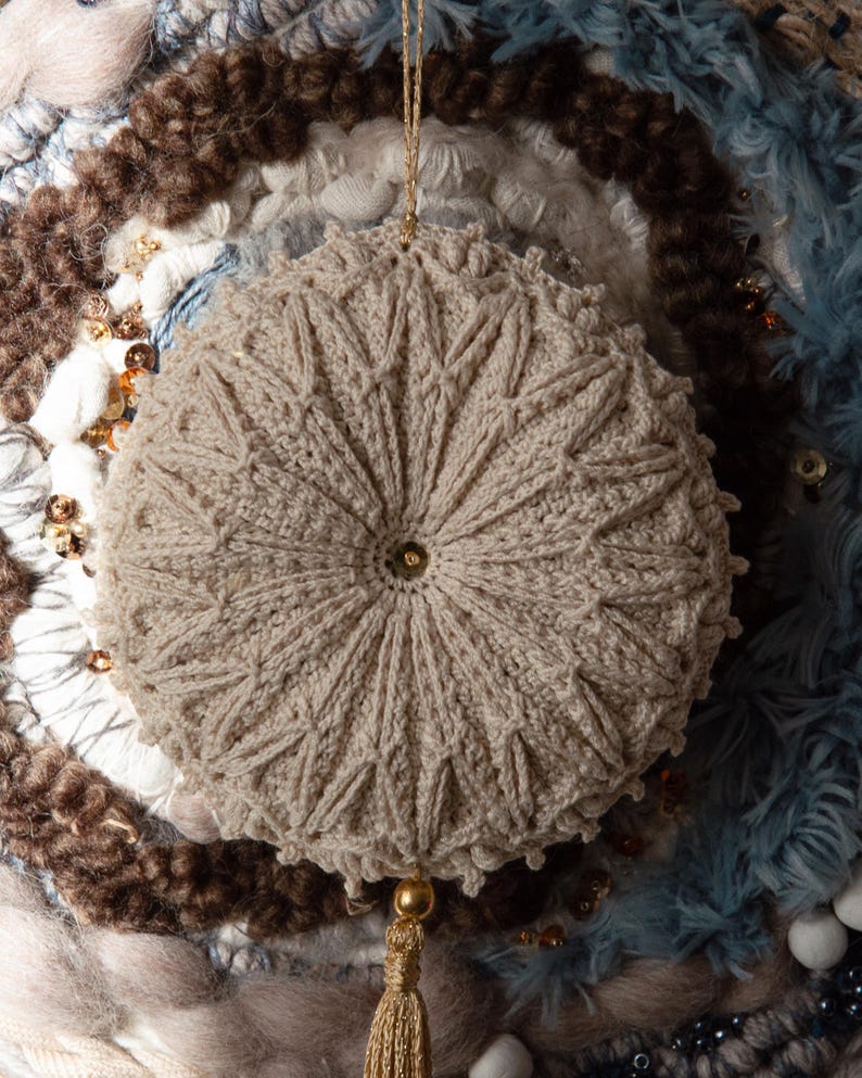 Puede incluir: Adorno beige tejido a ganchillo con dise&ntilde;o de estrella. El adorno redondo tiene una cuerda dorada para colgar y una borla. El fondo presenta varias texturas y colores, incluyendo azul, marr&oacute;n y blanco.