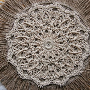 May include: A beige, circular, handmade doily with a detailed, openwork crochet design. The doily is centered on a round, fringed jute mat, creating a rustic, textured look. The background has a striped pattern.