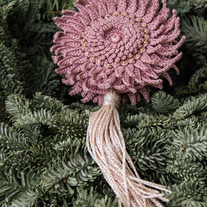 Puede incluir: Adorno floral de ganchillo rosa empolvado hecho a mano, adornado con cuentas doradas y una perla central. Cuenta con una borla beige claro brillante y se muestra sobre agujas de pino verdes. Un art&iacute;culo decorativo &uacute;nico.
