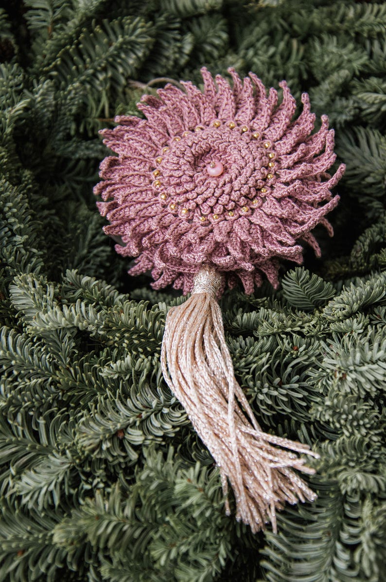 Puede incluir: Adorno floral de ganchillo rosa empolvado hecho a mano, adornado con cuentas doradas y una perla central. Cuenta con una borla beige claro brillante y se muestra sobre agujas de pino verdes. Un art&iacute;culo decorativo &uacute;nico.