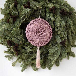 Puede incluir: Adorno de macram&eacute; rosa empolvado hecho a mano con detalles dorados y una borla a juego, colgado frente a una corona de hojas perennes verdes con pi&ntilde;as. El adorno redondo presenta un dise&ntilde;o floral detallado, ideal para la decoraci&oacute;n navide&ntilde;a.