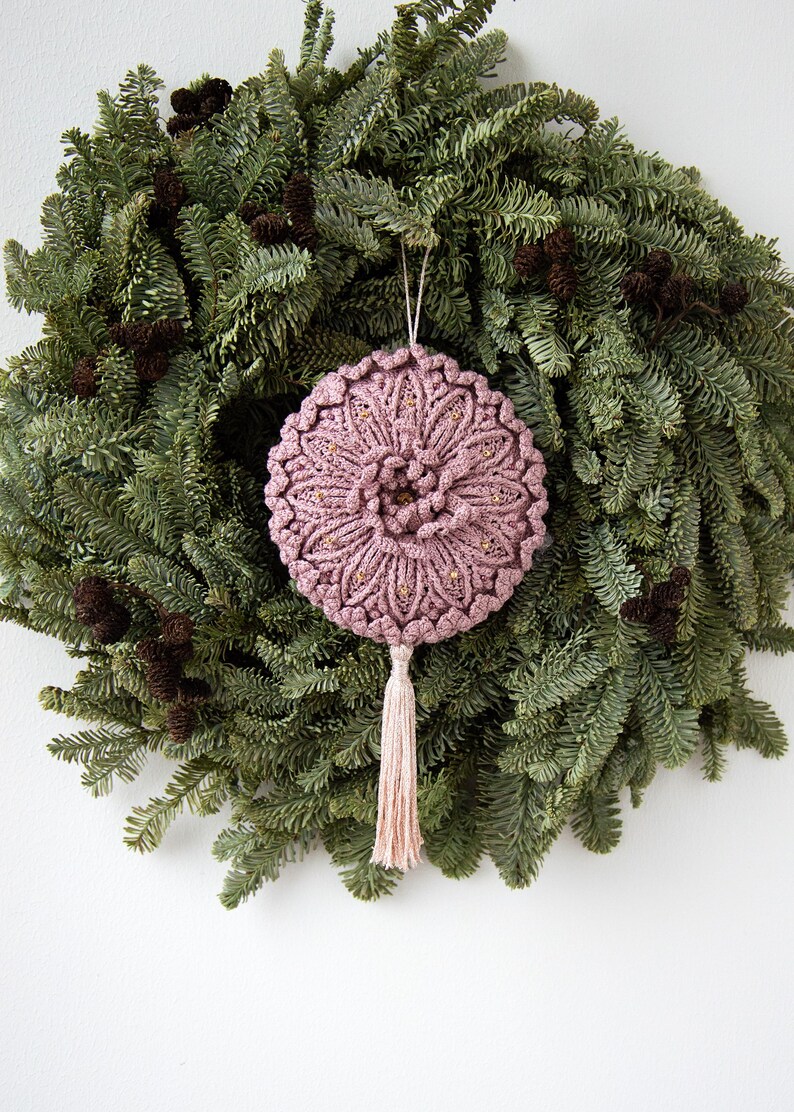 Puede incluir: Adorno de macram&eacute; rosa empolvado hecho a mano con detalles dorados y una borla a juego, colgado frente a una corona de hojas perennes verdes con pi&ntilde;as. El adorno redondo presenta un dise&ntilde;o floral detallado, ideal para la decoraci&oacute;n navide&ntilde;a.