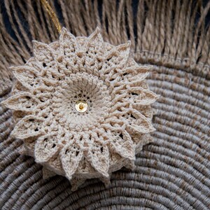 Puede incluir: Adorno floral beige tejido a ganchillo, con un centro de lentejuelas doradas. Se muestra sobre un salvamanteles circular tejido y un fondo tejido con flecos. Artesan&iacute;a detallada.