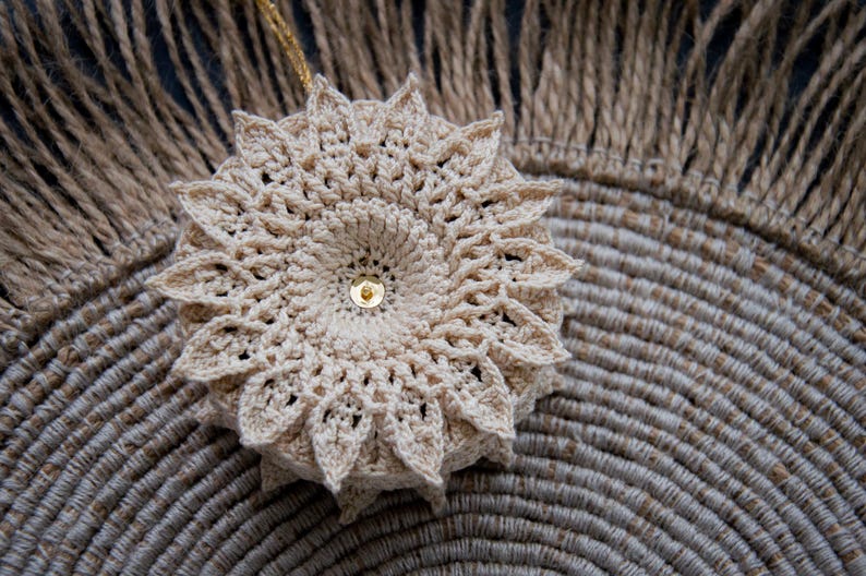 Puede incluir: Adorno floral beige tejido a ganchillo, con un centro de lentejuelas doradas. Se muestra sobre un salvamanteles circular tejido y un fondo tejido con flecos. Artesan&iacute;a detallada.