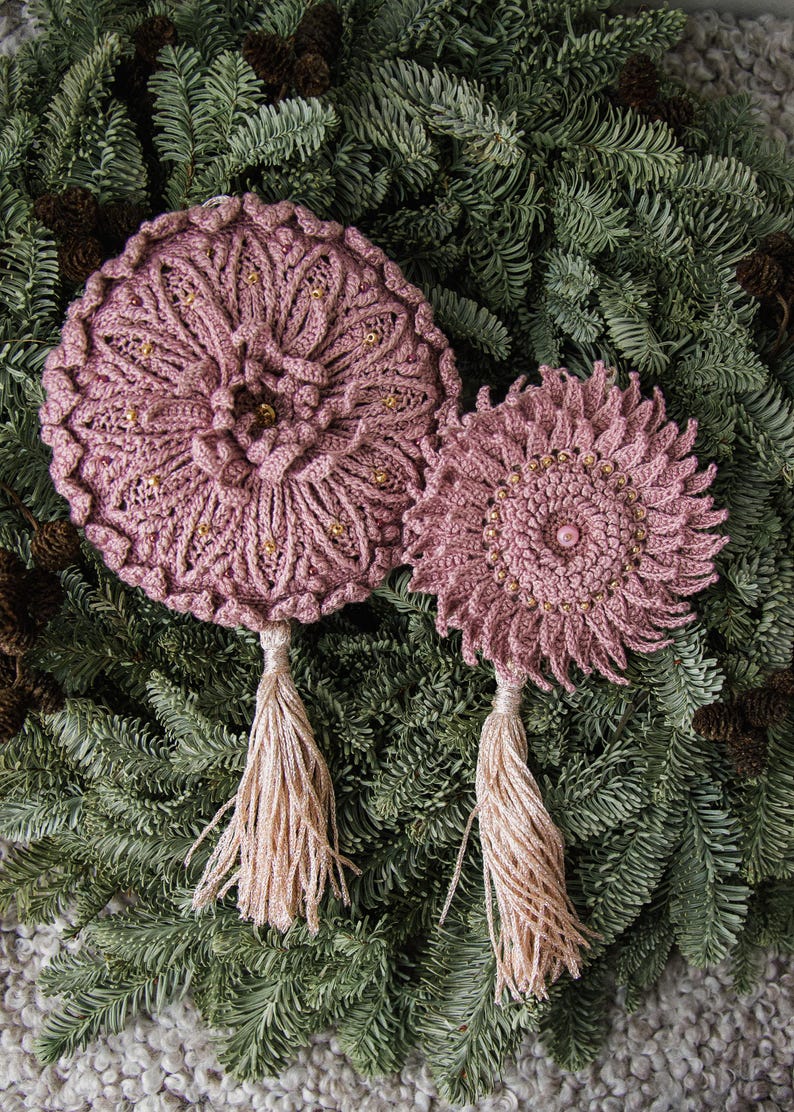Puede incluir: Dos adornos de ganchillo hechos a mano, de color rosa polvoriento, con detalles de cuentas doradas y largas borlas a juego. Los adornos se colocan sobre un fondo de agujas de pino verdes y peque&ntilde;as pi&ntilde;as.