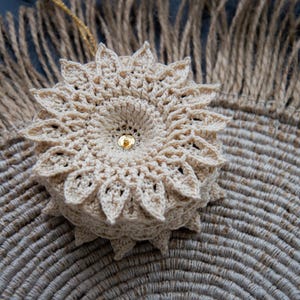 Puede incluir: Un adorno floral beige tejido a ganchillo con una cuenta central dorada, sobre un salvamanteles de yute tejido. El adorno tiene un dise&ntilde;o detallado en forma de p&eacute;talo. El fondo incluye un elemento de yute con flecos.