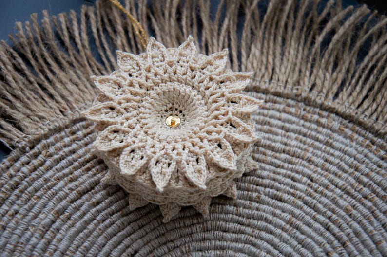 Puede incluir: Un adorno floral beige tejido a ganchillo con una cuenta central dorada, sobre un salvamanteles de yute tejido. El adorno tiene un dise&ntilde;o detallado en forma de p&eacute;talo. El fondo incluye un elemento de yute con flecos.